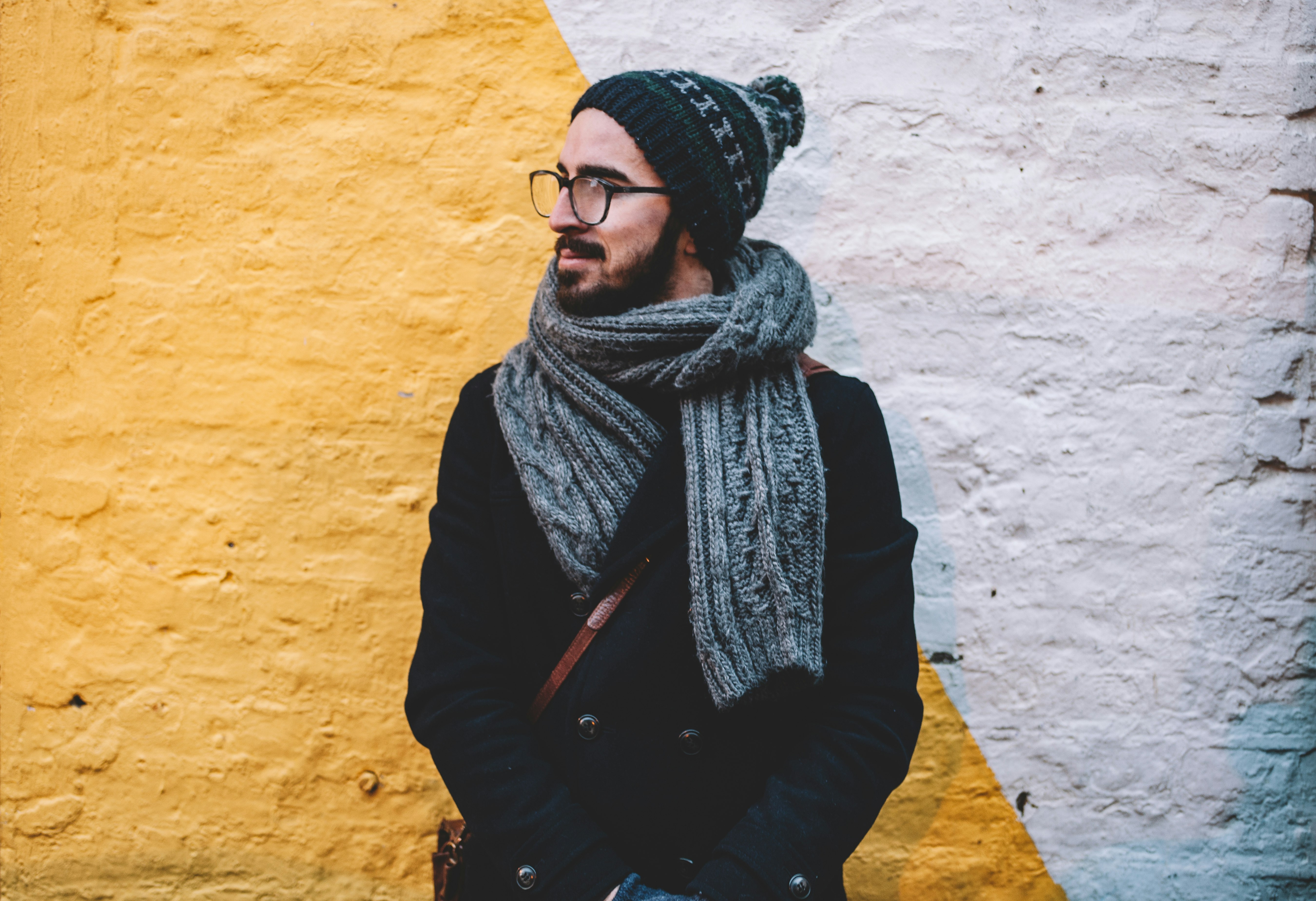 man in black coat with grey scarf standing on front of white and yellow painted wall